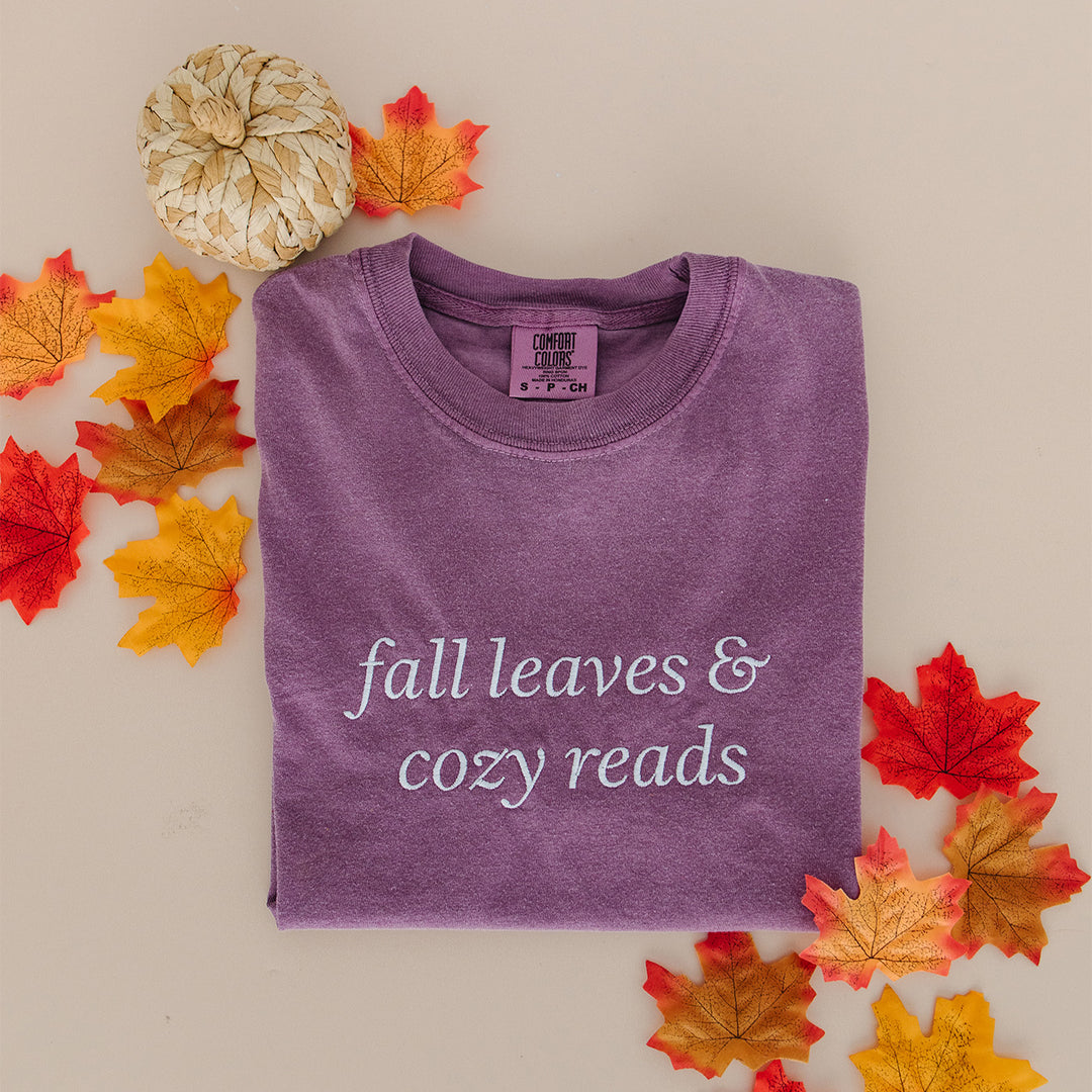 A folded purple t-shirt with the phrase 'fall leaves & cozy reads' in white embroidery, styled on a beige background with autumn leaves and a dried pumpkin.