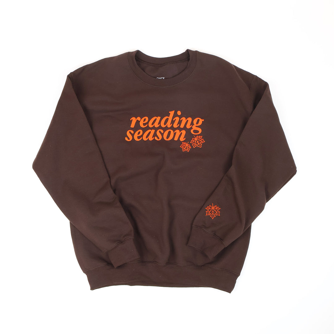 A dark brown crewneck sweatshirt with 'reading season' in orange embroidery across the chest, accompanied by two small orange leaves graphics. A small orange leaf graphic is also embroidered on the left sleeve cuff.
