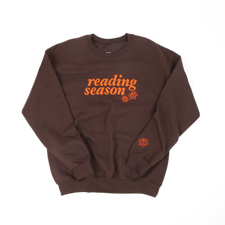 A dark brown crewneck sweatshirt with 'reading season' in orange embroidery across the chest, accompanied by two small orange leaves graphics. A small orange leaf graphic is also embroidered on the left sleeve cuff.