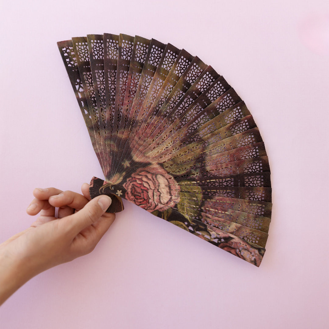 A hand holds out a custom rose-patterned, brown wood handheld fan