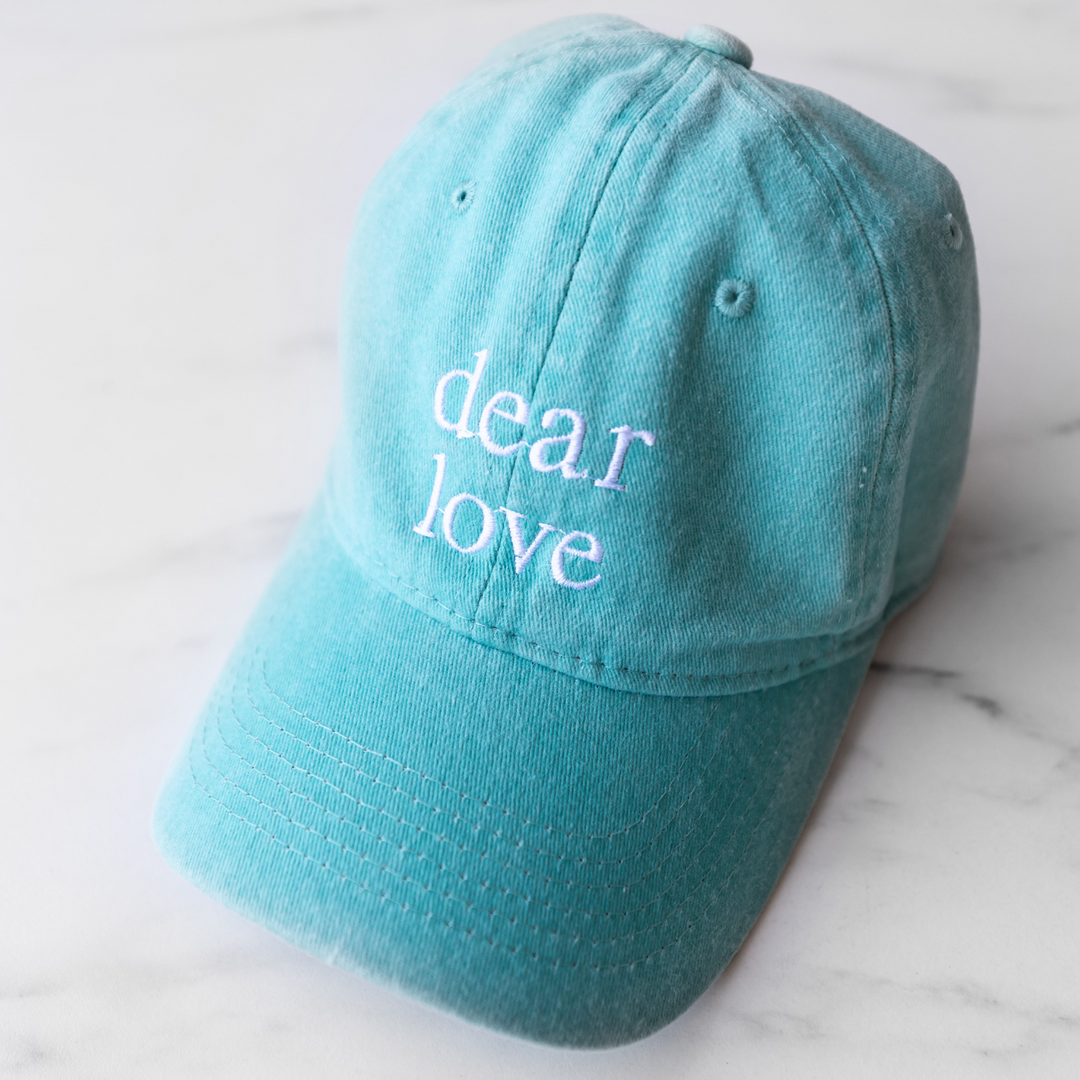 A light blue baseball hat featuring the words "Dear Love" in white embroidery thread.