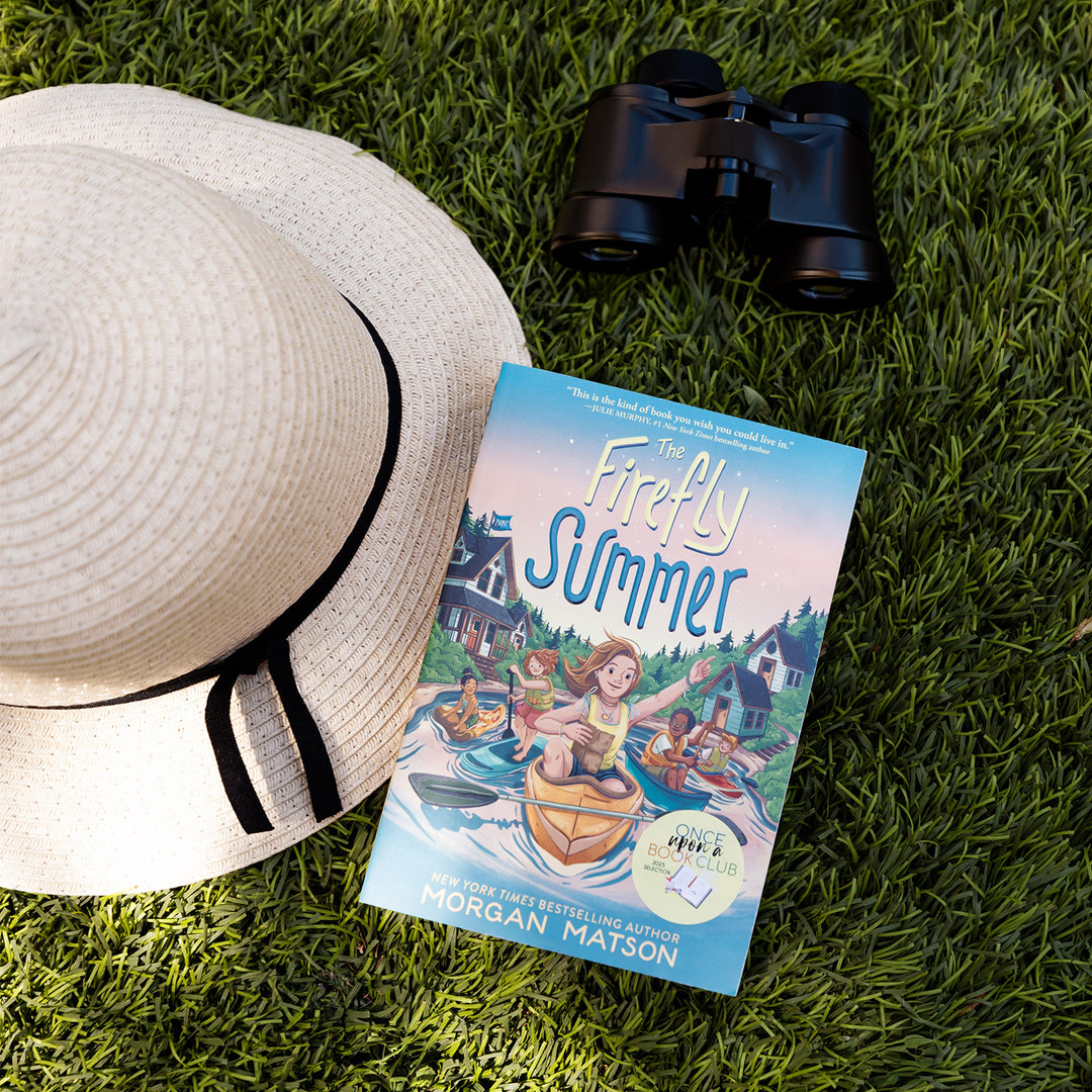 A paperback copy of The Firefly Summer by Morgan Matson sits on grass. Binoculars and a hat surround the book.