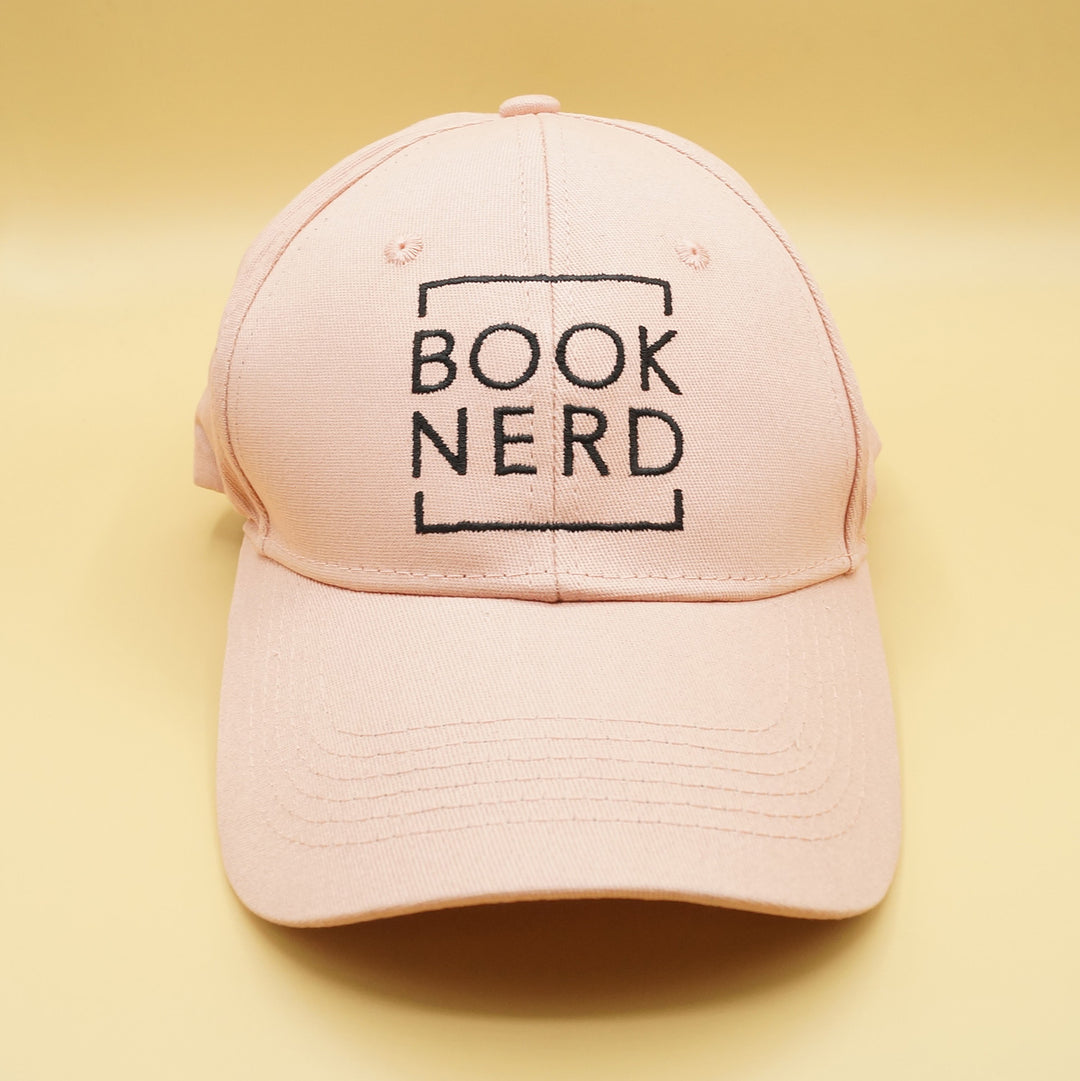 a pale pink baseball cap that says BOOK NERD in black writing on the front