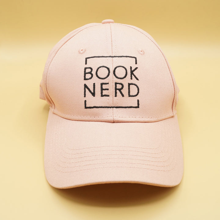 a pale pink baseball cap that says BOOK NERD in black writing on the front