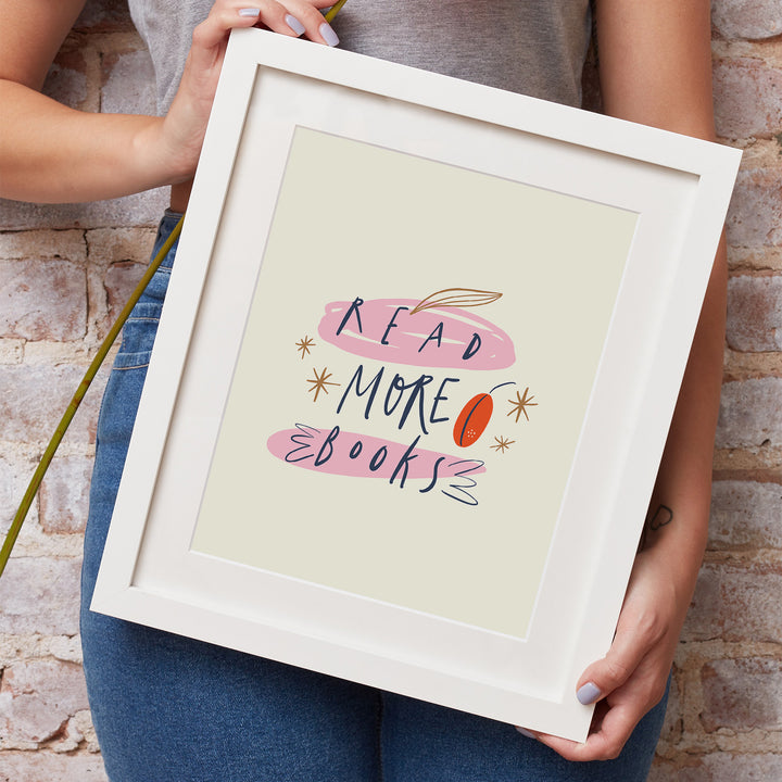 A white woman's hands hold a white matted frame showcasing the "Read More Books" art print. She is wearing jeans and a grey shirt and leaning against a brick wall.