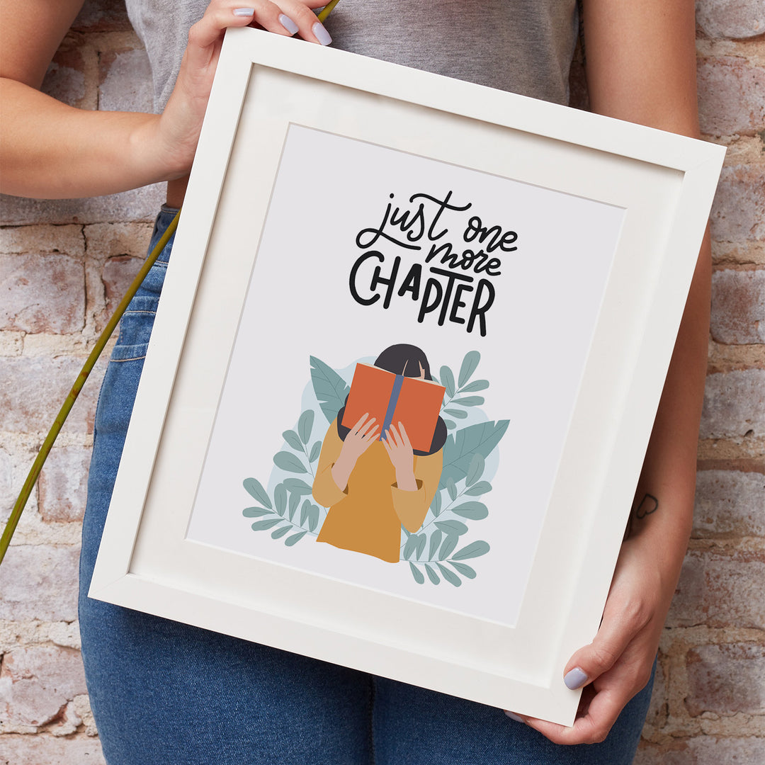 A white woman's hands hold a white matted frame showcasing the "One More Chapter" art print. She is wearing jeans and a grey shirt and leaning against a brick wall.