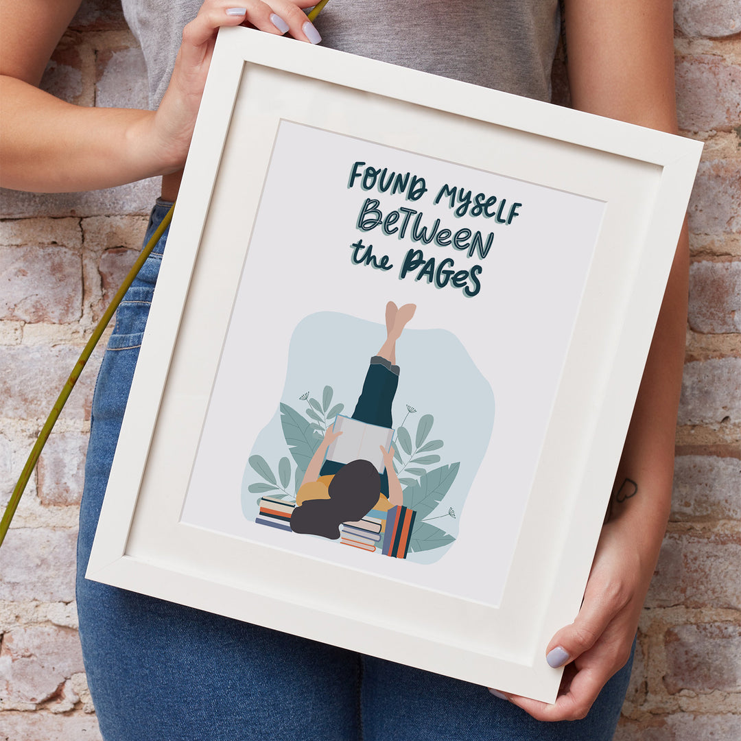 A white woman's hands hold a white matted frame showcasing the "Between the Pages" art print. She is wearing jeans and a grey shirt and leaning against a brick wall.