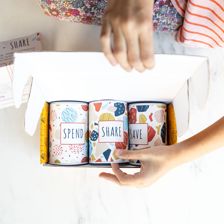 A white hand pulls the "Share" tin from its box with the "Spend" and "Save" tin.