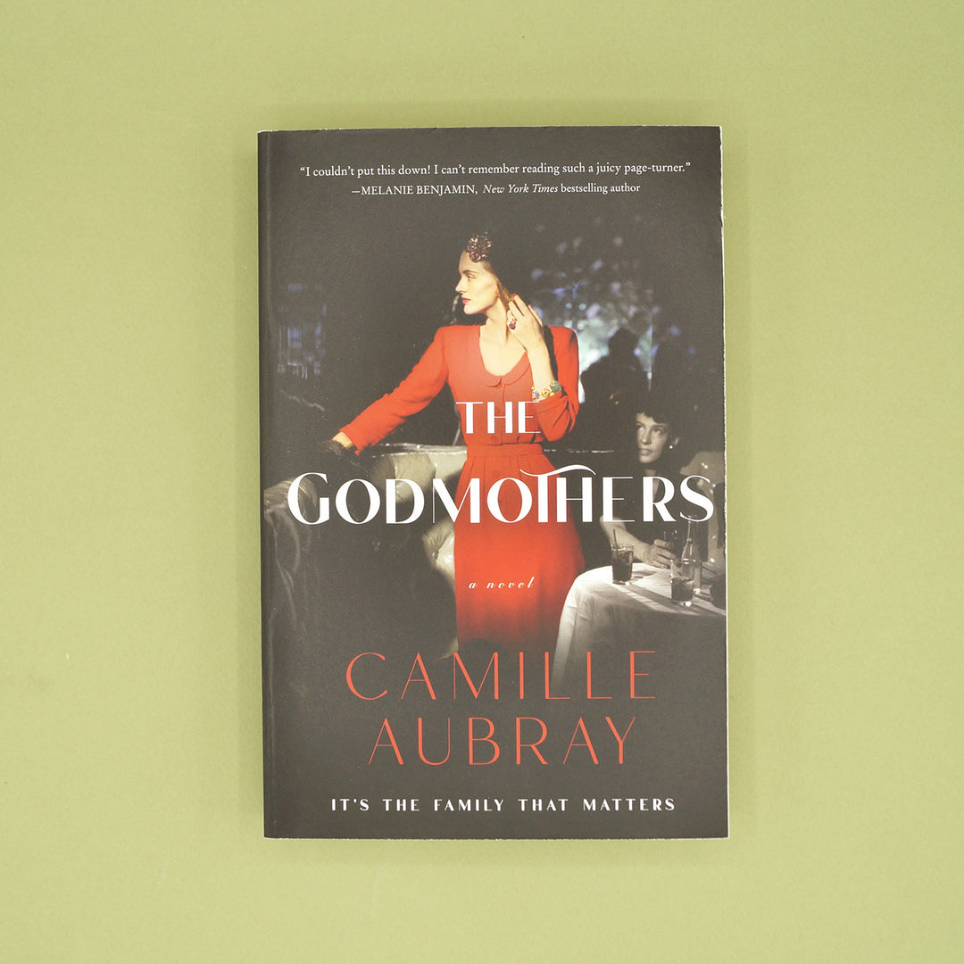 A paperback copy of The Godmothers by Camille Aubray sits on a green background.