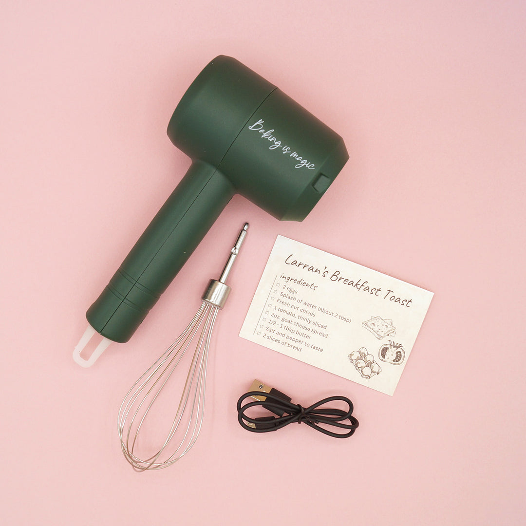 a wireless, dark green hand mixer with whisk attachment, charging cables, and a recipe card sits on a pink background. The mixer has the words "Baking is Magic" printed on the top barrel.