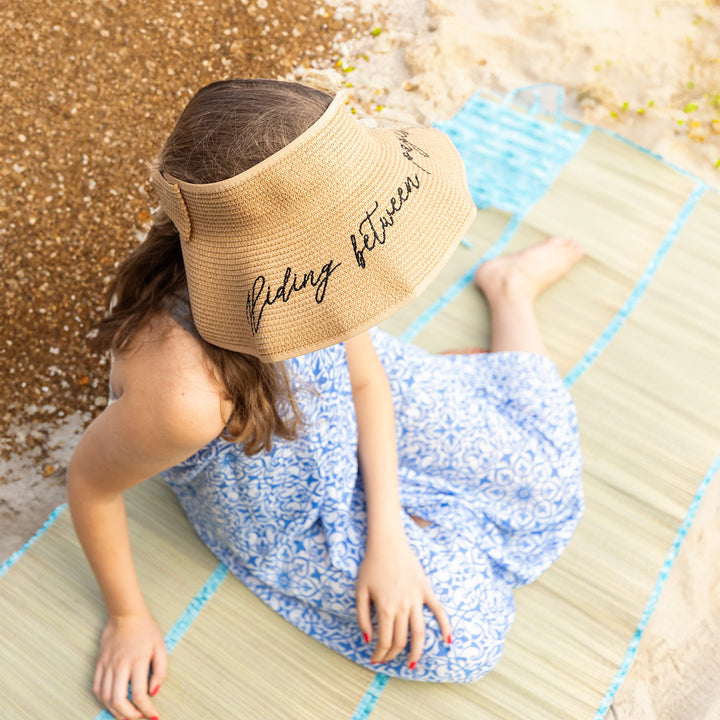 A girl sits on an oversized tatami beach mat with the quote: The air still tastes of salt and memory. The quote is on a square blue section of the mat. She wears an open top, sand-toned sun hat featuring an embroidered quote on the brim of the hat: hiding between pages of books