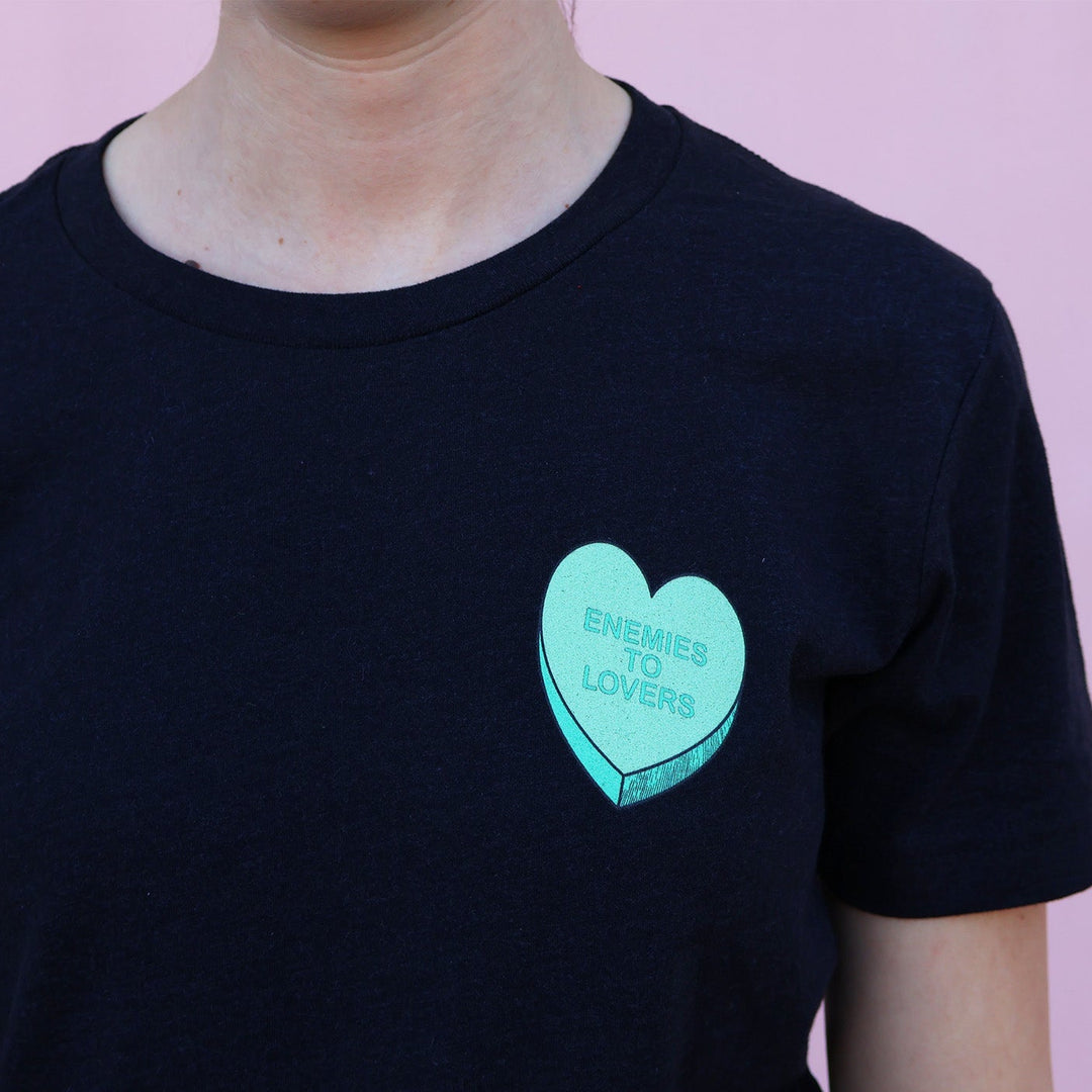 Black t-shirt with a green heart-shaped candy design and 'Enemies to Lovers' text on a pink background.