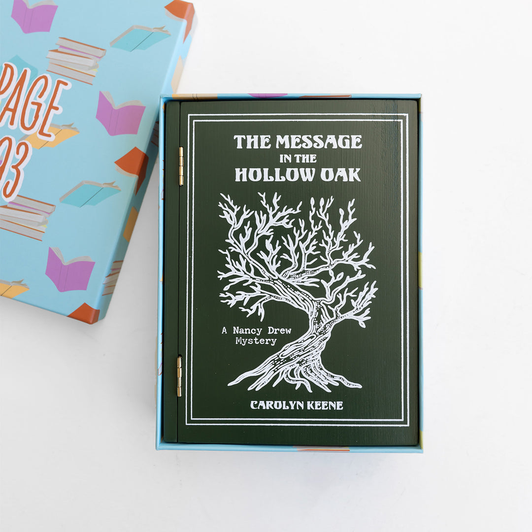 An open box with a dark green book-shaped safe. In white text, the front says 'The Message in the Hollow Oak' 'A Nancy Drew Mystery' 'Carolyn Keene'. with an image of a white oak tree.