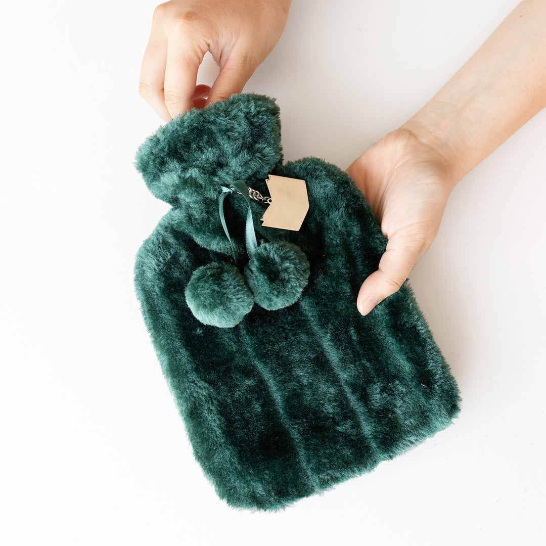 Two hands hold a dark green fuzzy hot water bottle with a book-shaped keychain tied to it.