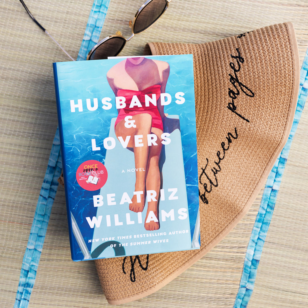 A hardcover copy of Husbands & Lovers by Beatriz Williams sits on a beach mat surrounded by sunglasses and a beach hat.