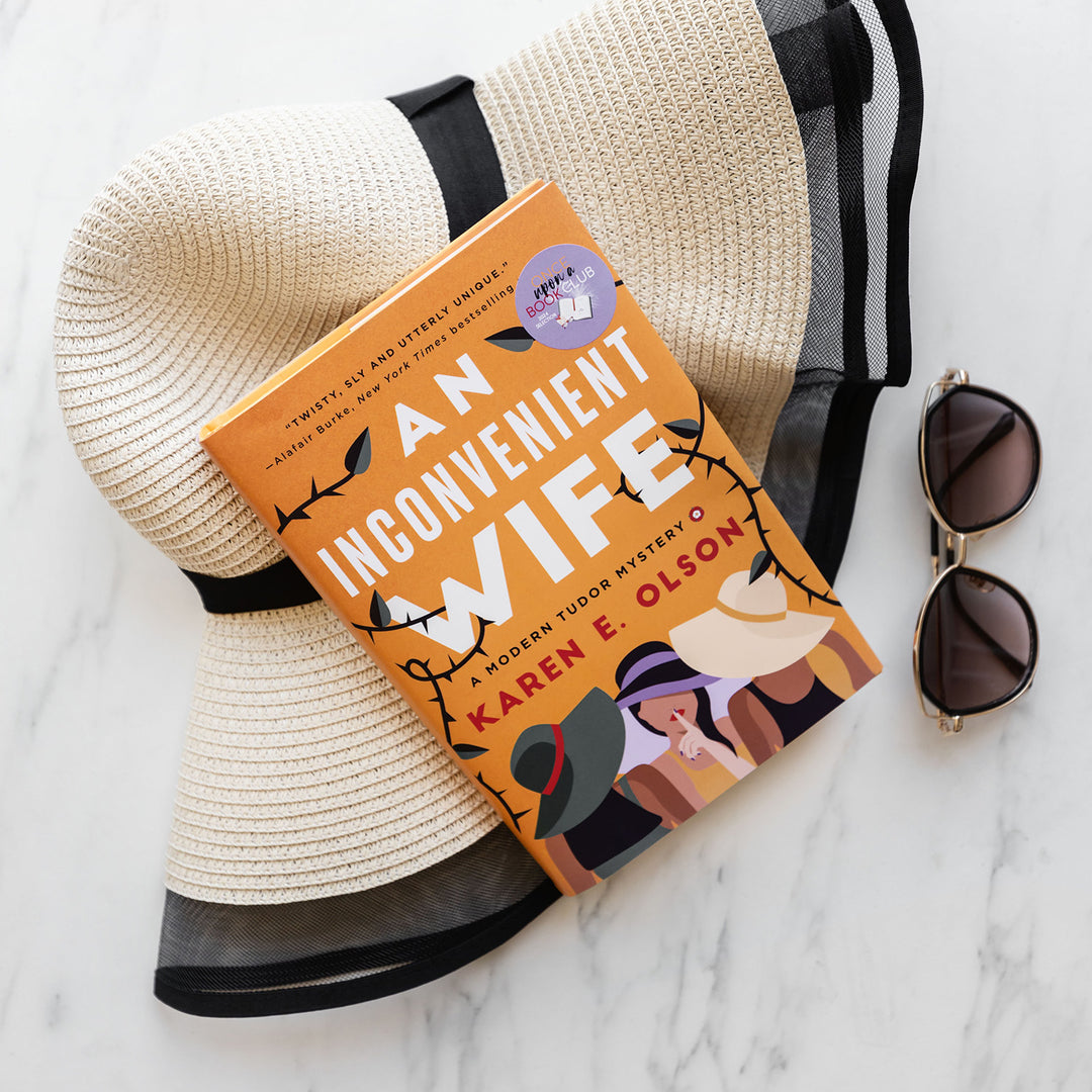 a hardcover copy of AN INCONVENIENT WIFE by Karen E. Olson rests on a sunhat and a pair of sunglasses sits next to it.