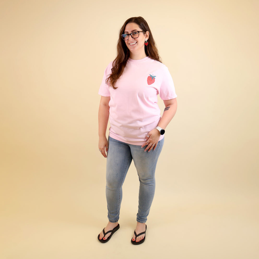 Woman wearing a pink t-shirt with a strawberry designs and text that says 'Reading is my Jam' on a beige background