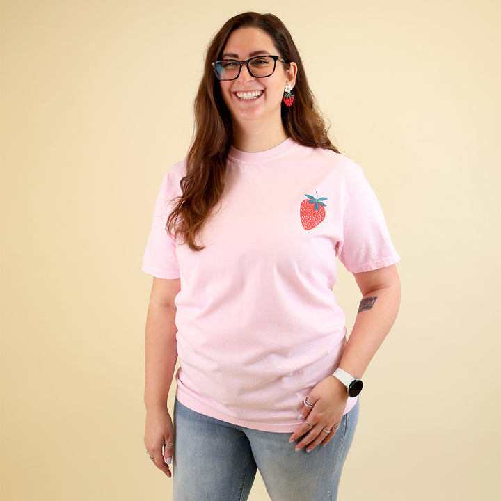 Woman wearing a pink t-shirt with a strawberry designs and text that says 'Reading is my Jam' on a beige background
