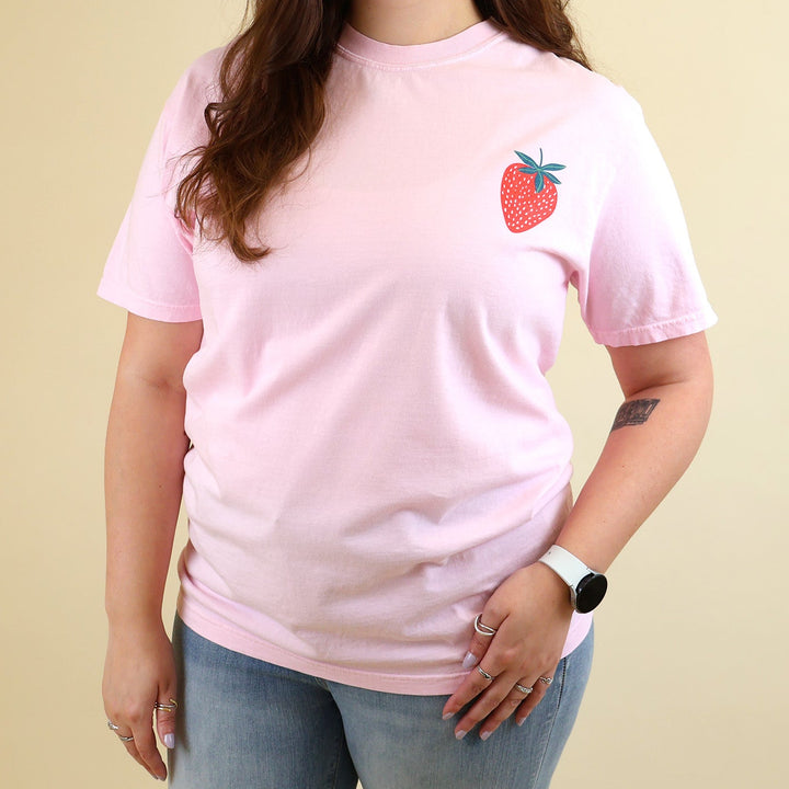 Woman wearing a pink t-shirt with a strawberry designs and text that says 'Reading is my Jam' on a beige background
