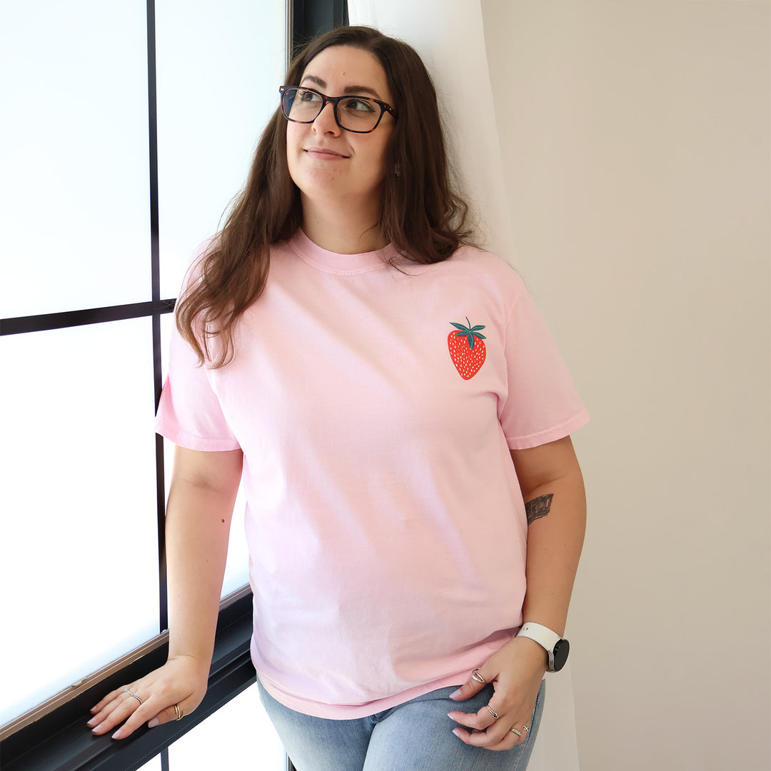 Woman wearing a pink t-shirt with a strawberry designs and text that says 'Reading is my Jam' against a window.