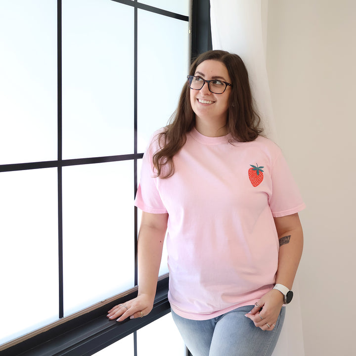 Woman wearing a pink t-shirt with a strawberry designs and text that says 'Reading is my Jam' against a window.