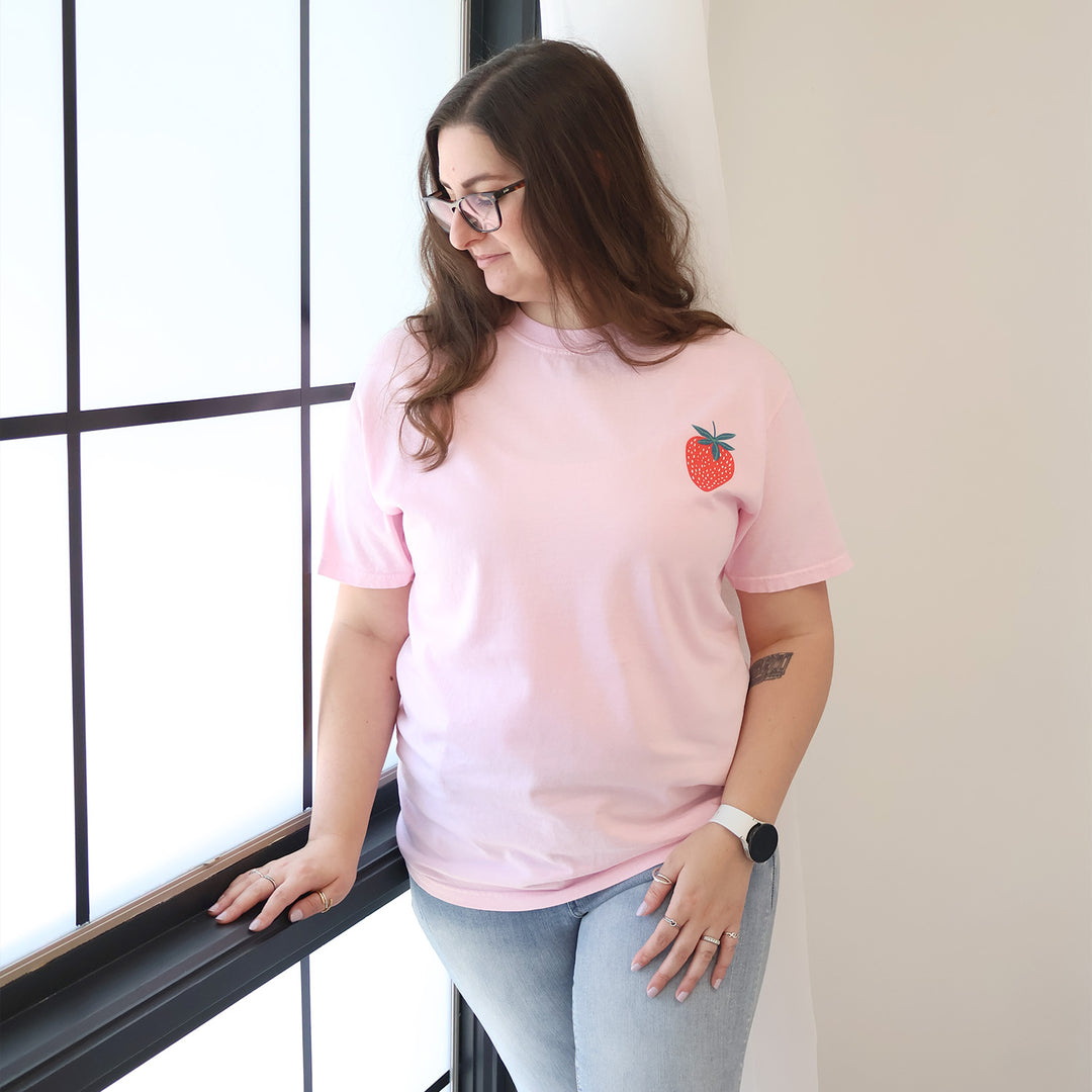 Woman wearing a pink t-shirt with a strawberry designs and text that says 'Reading is my Jam' stands near a window.