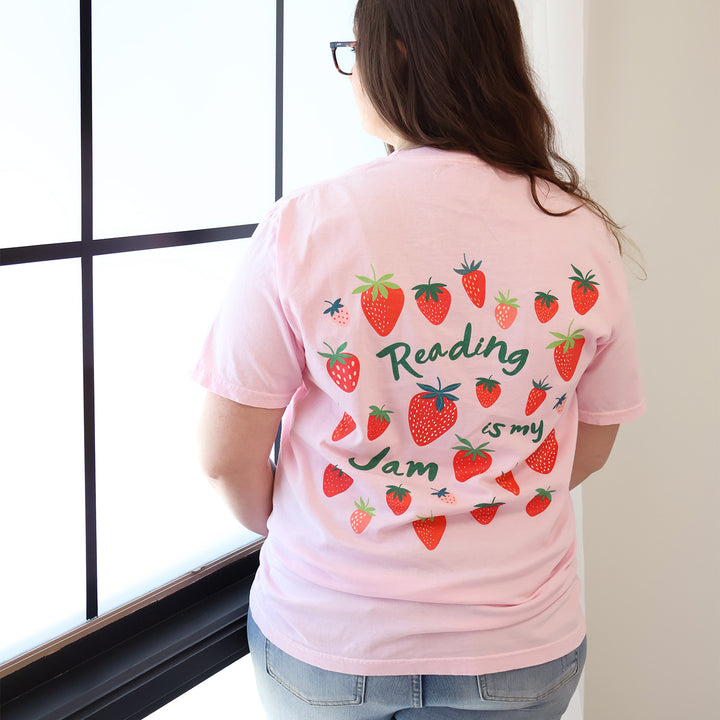 Woman wearing a pink t-shirt with a strawberry designs and text that says 'Reading is my Jam' stands near a window.