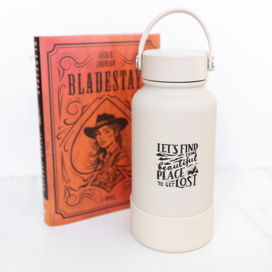 A light taupe colored metal water bottle with handle lid sits on a white background in front of the OUABC special edition of Bladestay by Jackie Johnson. The water bottle is printed with clouds and mountains and the words "Let's find some beautiful place to get lost."