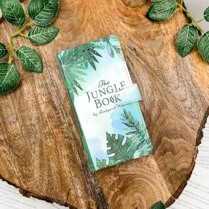 Book-shaped phone case titled 'The Jungle Book' with a jungle-themed cover on a wooden surface with leaves.
