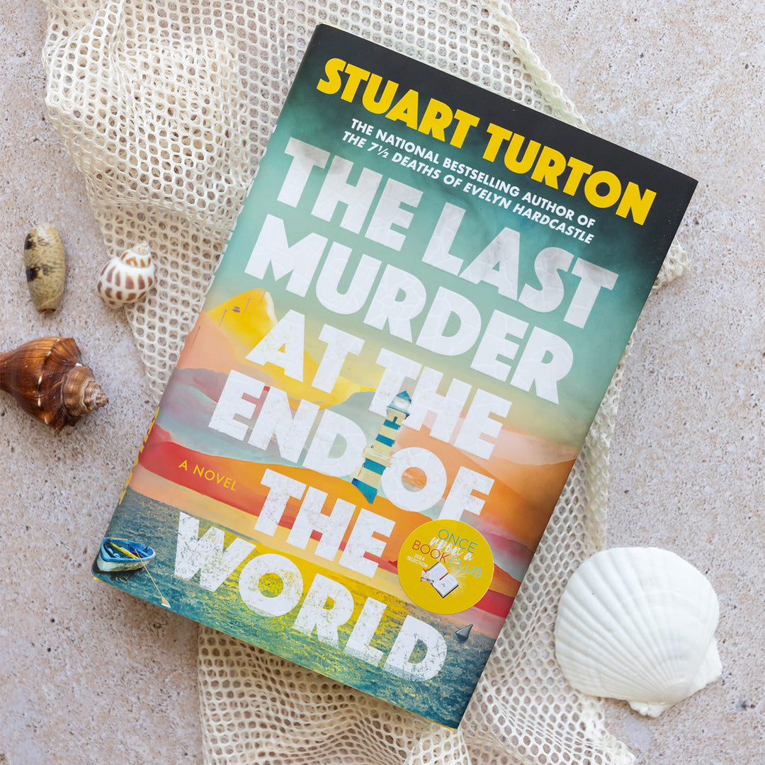 a hardcover copy of The Last Murder at the End of the World by Stuart Turton surrounded by seashells and sand.