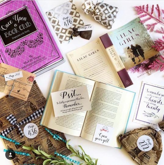 A hardcover copy of Lilac Girls by Martha Hall Kelly sits open. Surrounded it are wrapped gifts labeled with page numbers. In the upper left corner of the image is a pink Once Upon a Book Club box.