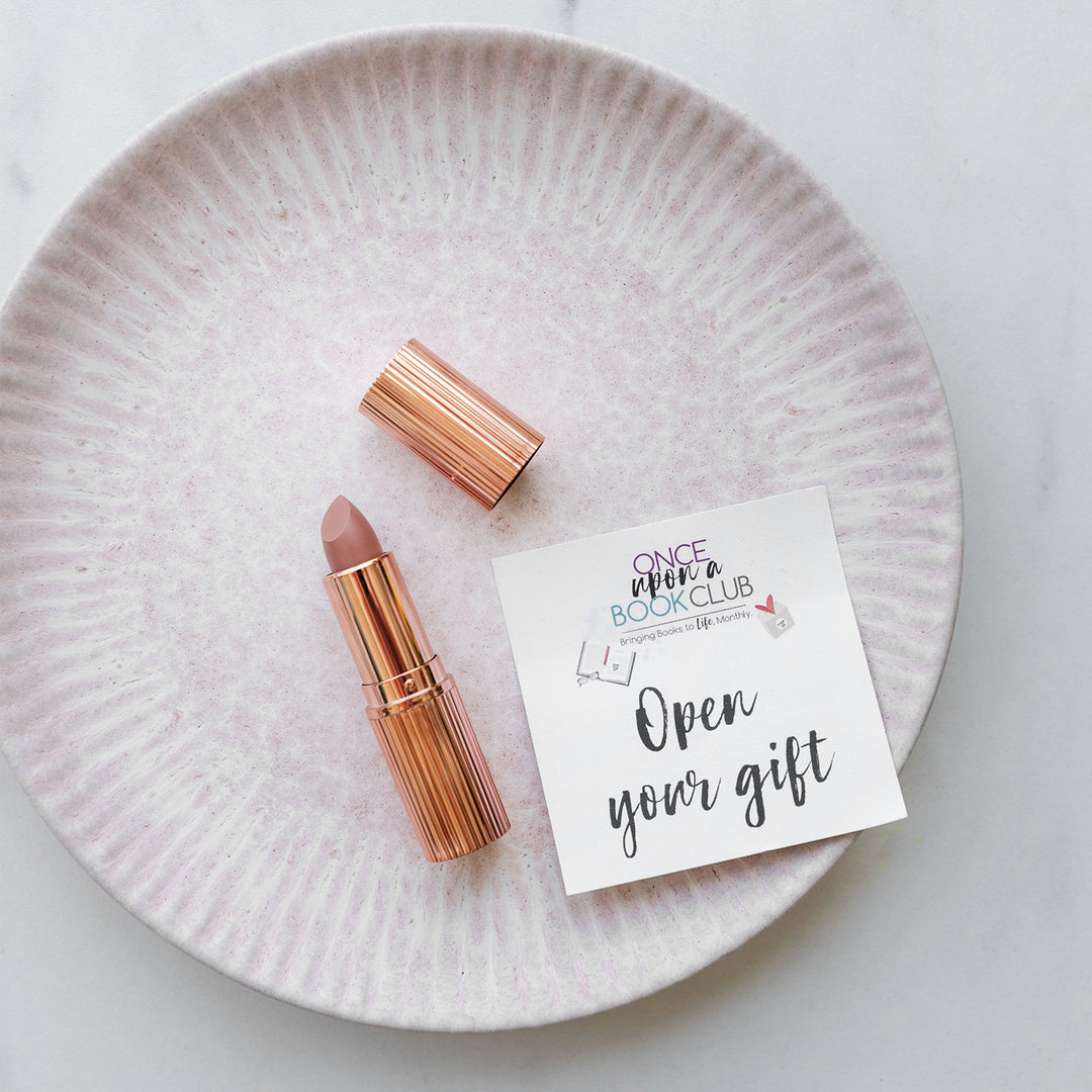 A lipstick tube is sitting open on a white plate and shows a nude color lipstick. The gold cap is next to it along with an open your gift sticker.