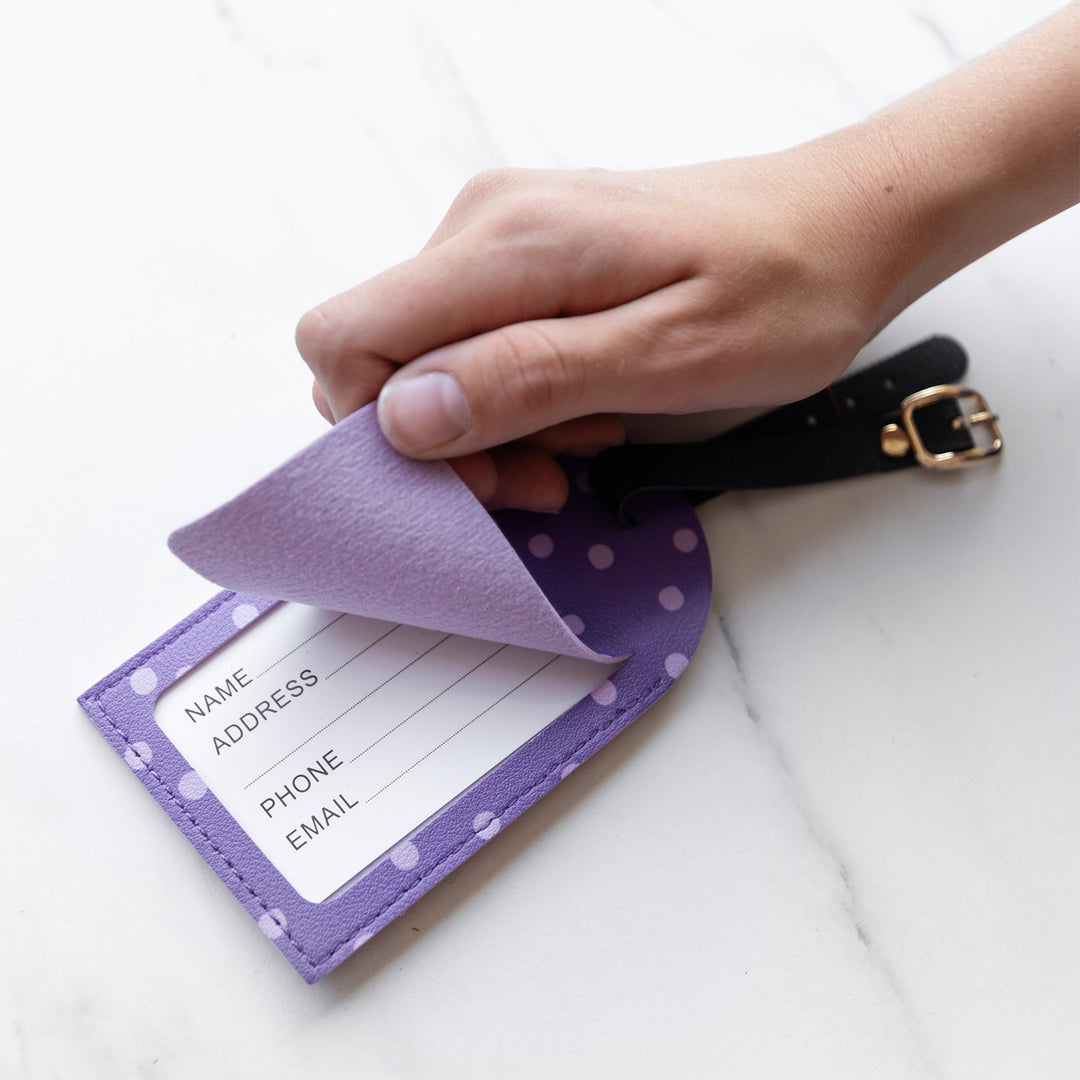 A purple luggage tag with light purple polka dots and a black strap. A white hand lifts up the flap to show space to write your name, address, phone, and email underneath.