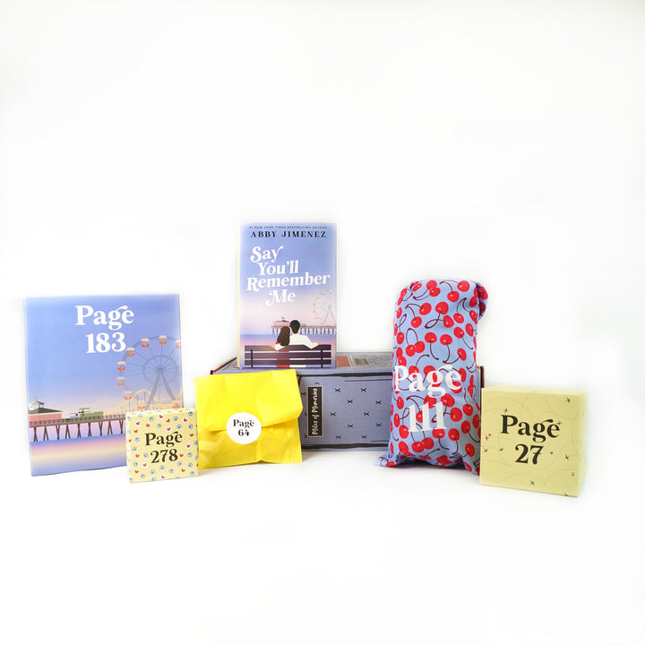 A hardcover, exclusive edition of "Say You'll Remember Me" by Abby Jimenez stands on a purple box surrounded by wrapped gifts labeled with page numbers.