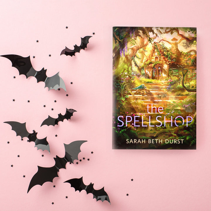 A hardcover copy of The Spellshop by Sarah Beth Durst sits on a pink background. Next to it are black cut-outs of bats and tiny black stars.