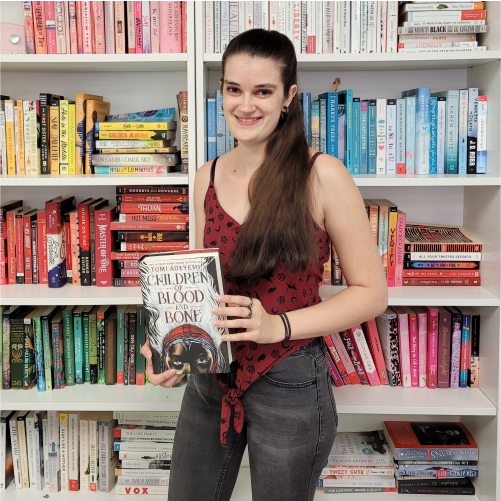 A woman wearing a red patterned top and jeans smiles while holding the book 