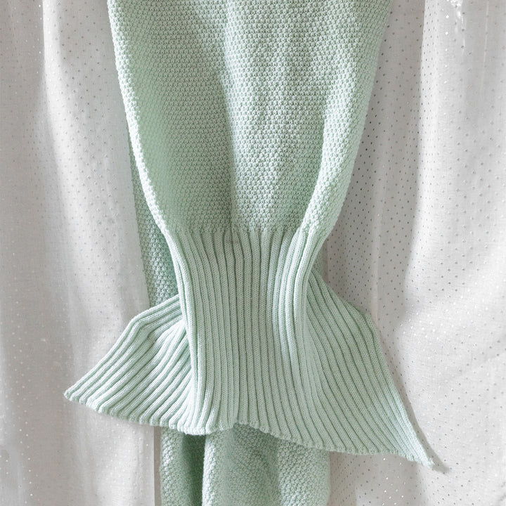 a seafoam green, knit mermaid tail blanket lays on a white background