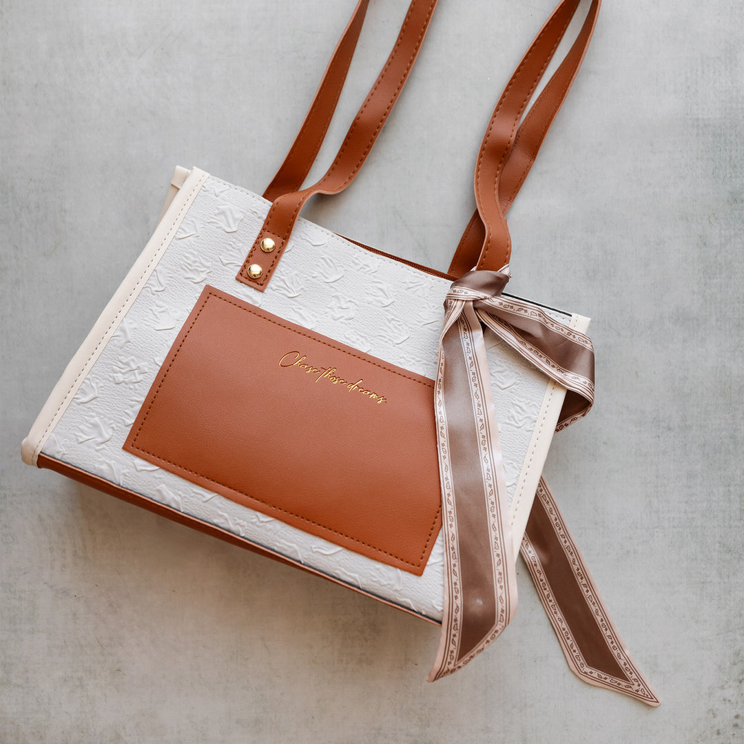 A brown and white purse that says Chase Those Dreams in small gold lettering on the front. A light brown silk tie is tied to one strap of the purse.