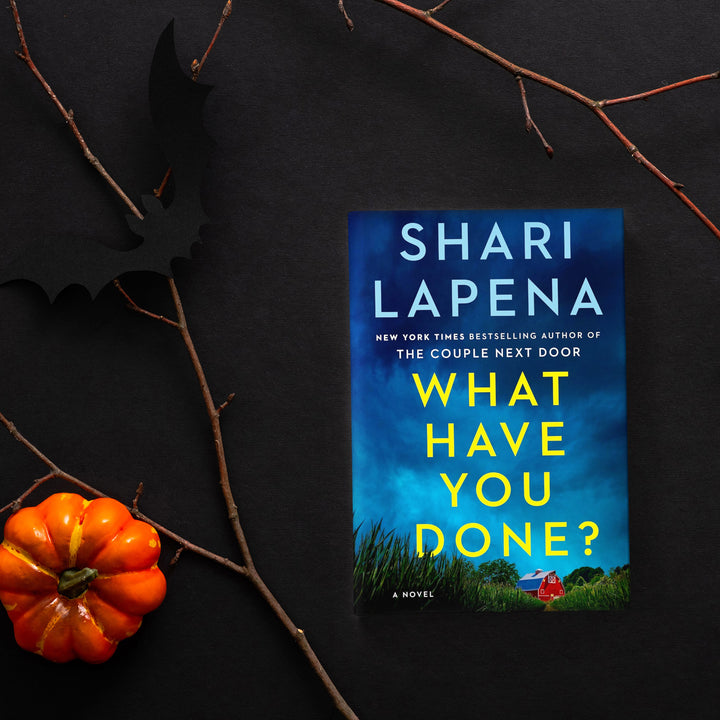 A hardcover copy of What Have You Done? by Shari Lapena sits on a black background. Surrounding the book are tree branches, a small orange pumpkin, and a black cut-out of a bat.