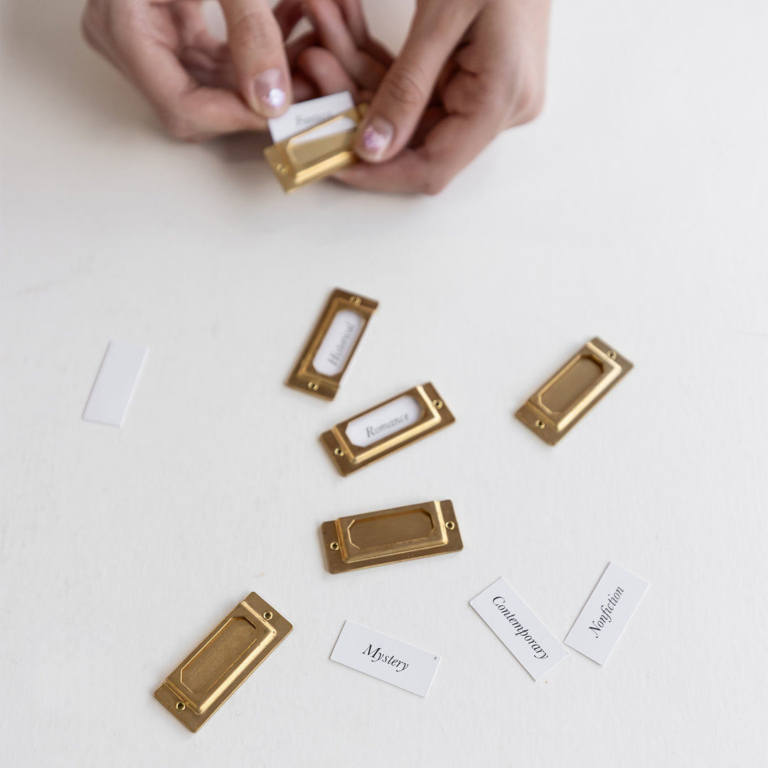 Small gold labels lay scattered on the table and a hand holds one while putting a slip of paper in the label. There are other slips of paper next to the labels that say Historical, Romance, Mystery, Contemporary, and Nonfiction.