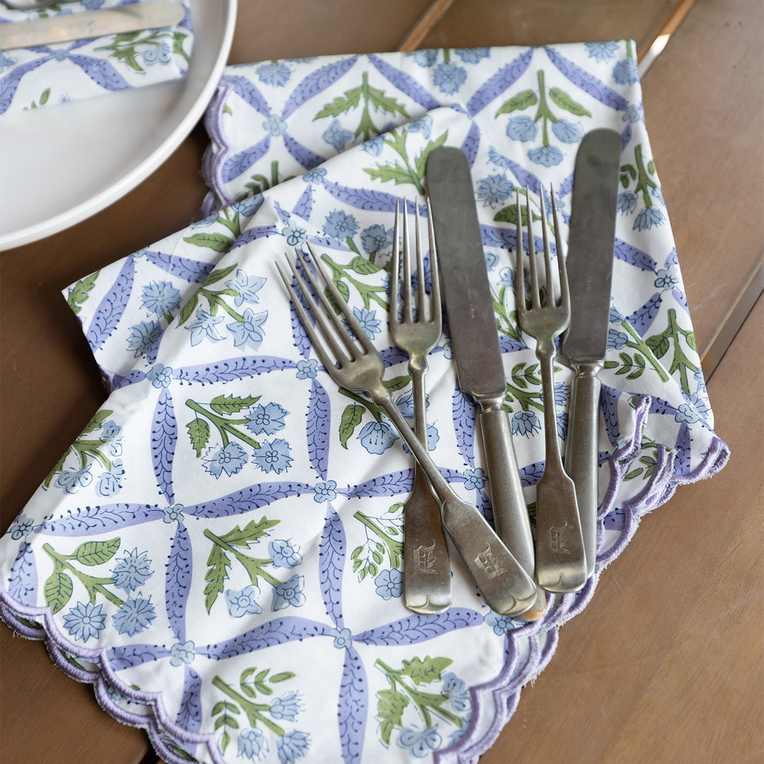 Three purple floral and white napkins are stacked on a brown wooden table with forks and knives laying on top of them. 