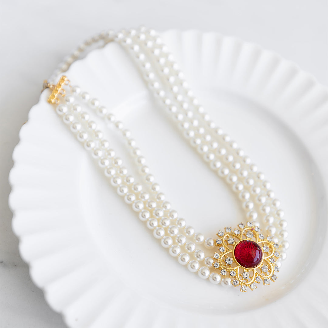 A three-strand pearl necklace with a gold and diamond flower-shaped piece with a ruby at the center.