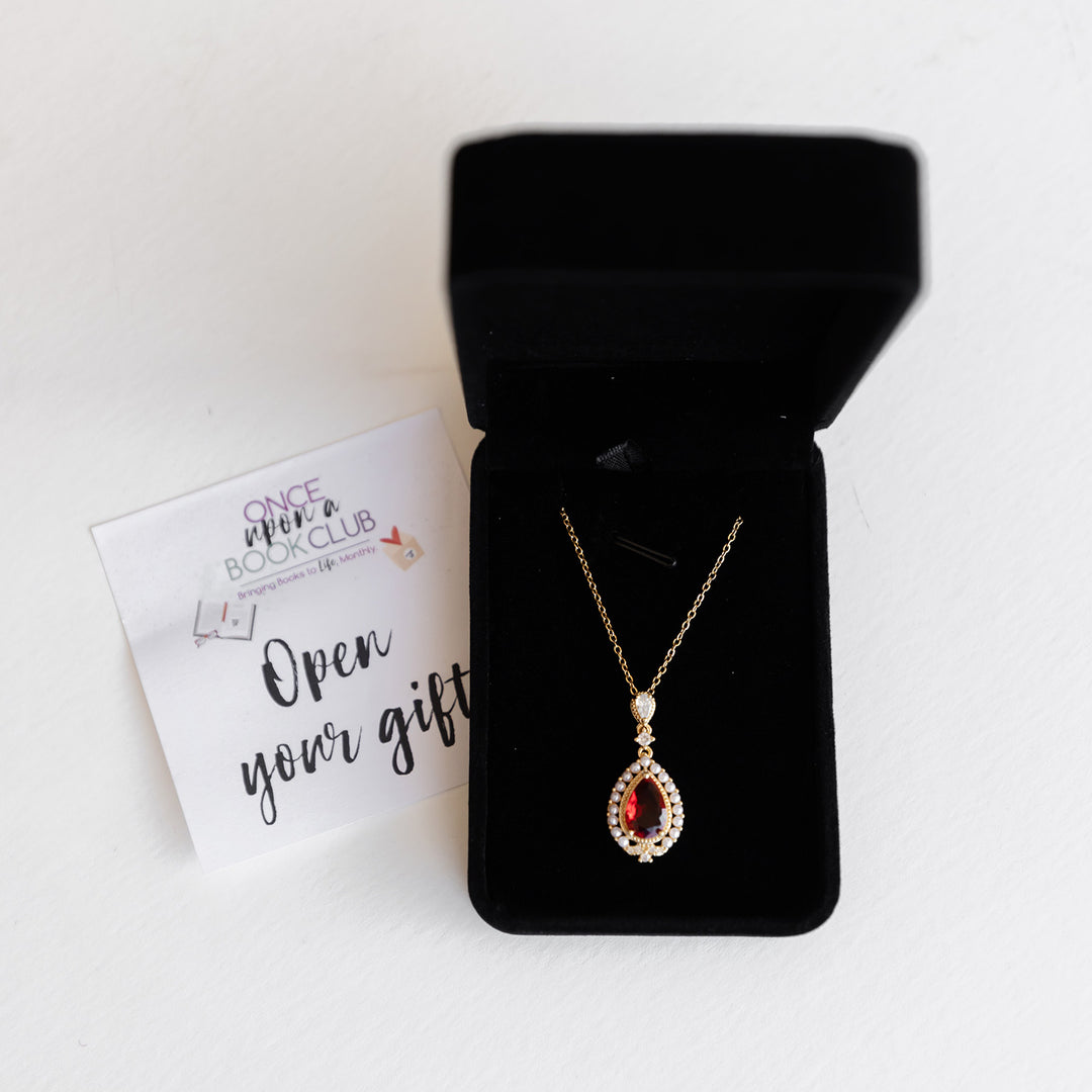 An open black jewelry box with a ruby necklace surrounded by small pearls on a gold chain. The box is next to an Open Your Gift sticker