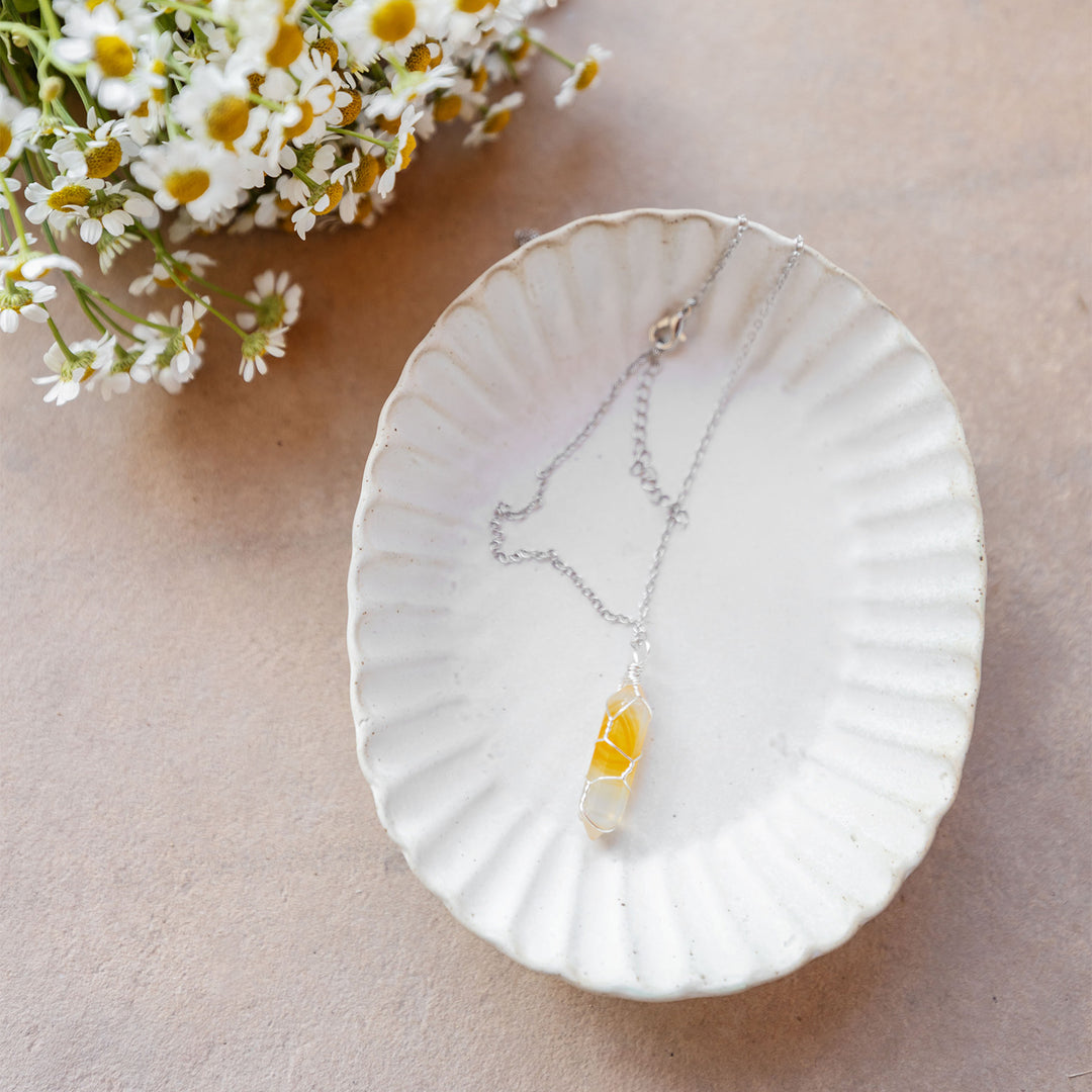 A white dish sits on a beige background. The dish contains a silver chain with a yellow and clear stone wrapped in silver wire. Daisies are in the upper left corner.