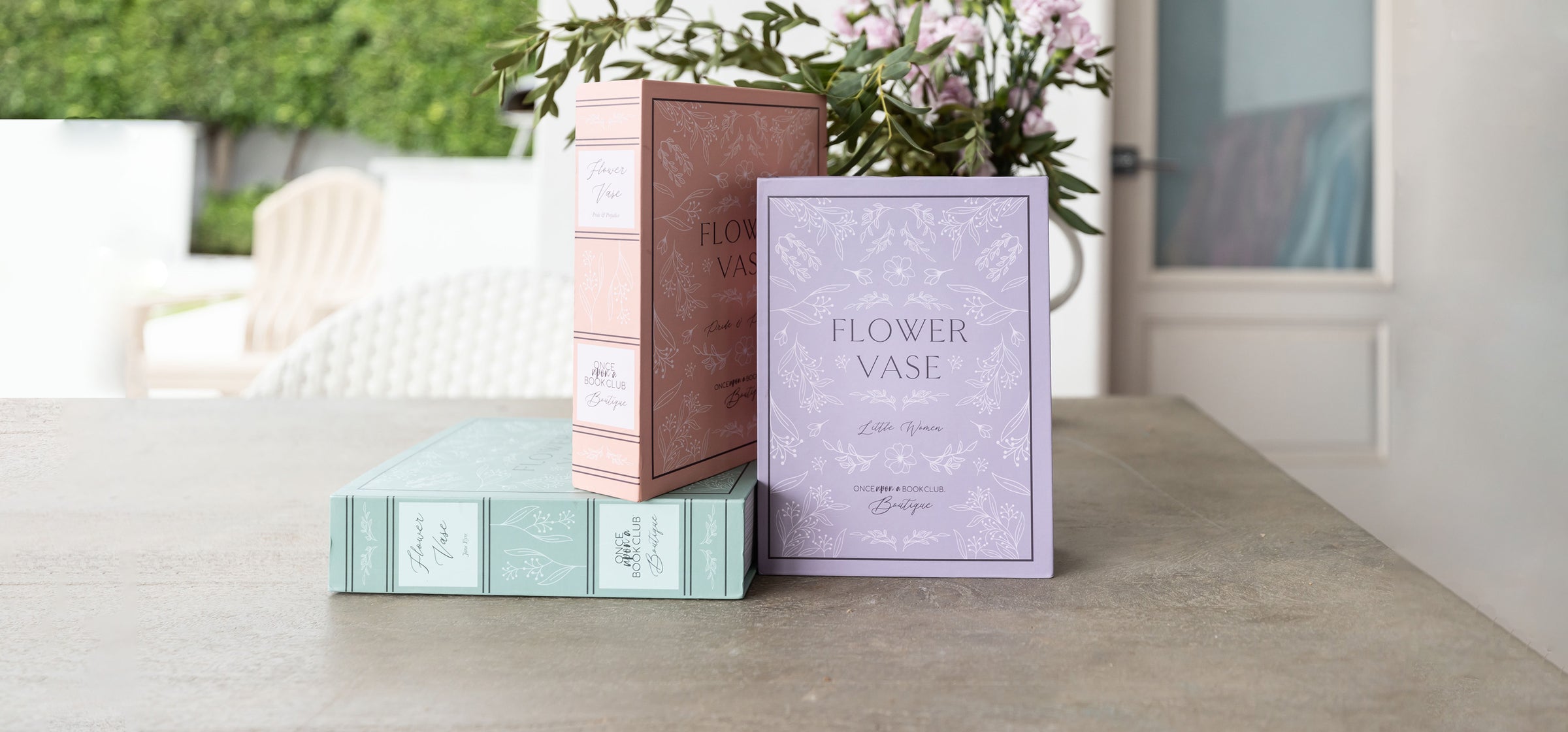 A set of three different colored boxes sit on a countertop with flowers behind them. All three boxes read "Flower Vase" along with the book title that inspired each vase design and "Once Upon a Book Club Boutique" is written across the bottom of each box.