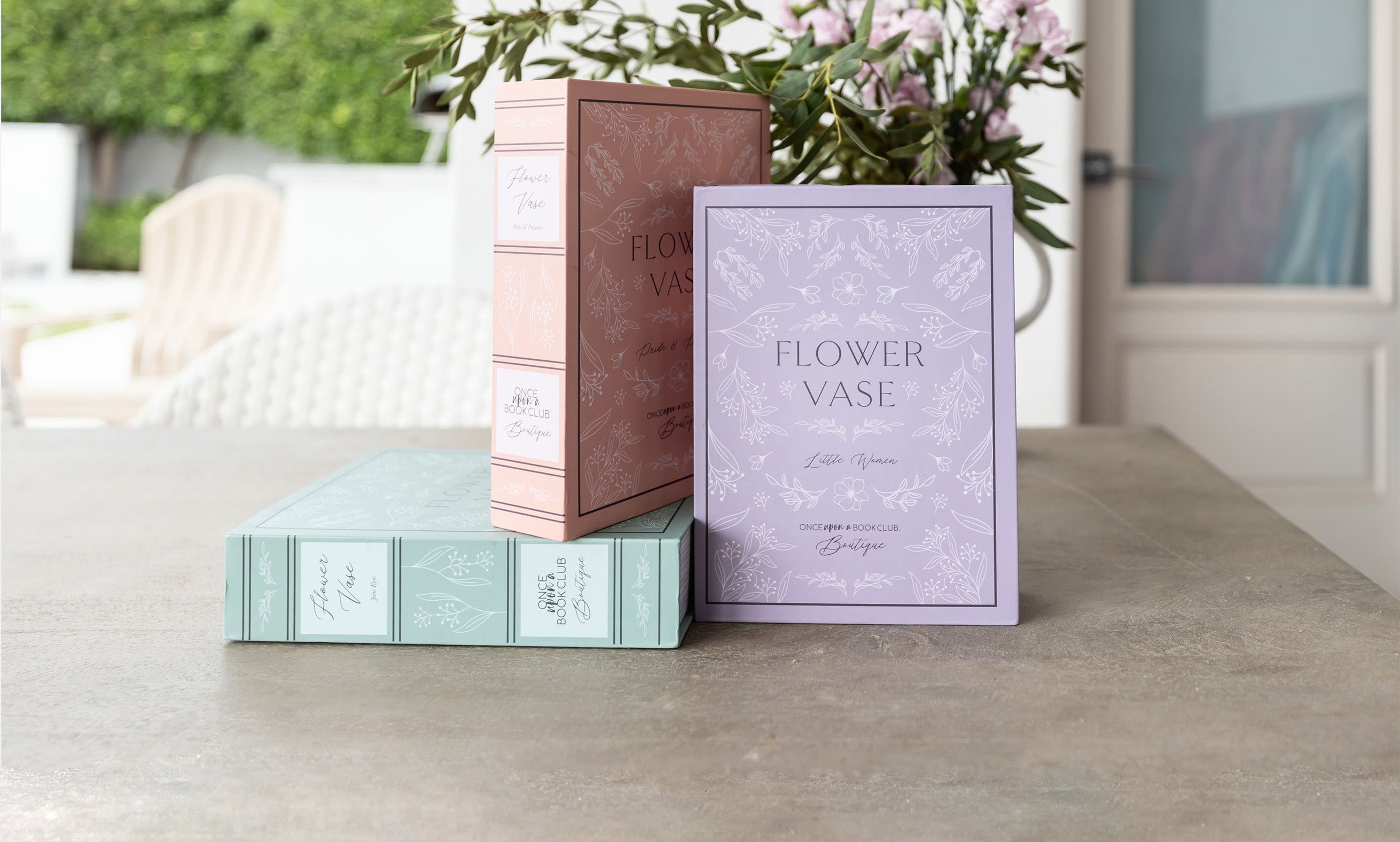 A set of three different colored boxes sit on a countertop with flowers behind them. All three boxes read "Flower Vase" along with the book title that inspired each vase design and "Once Upon a Book Club Boutique" is written across the bottom of each box.