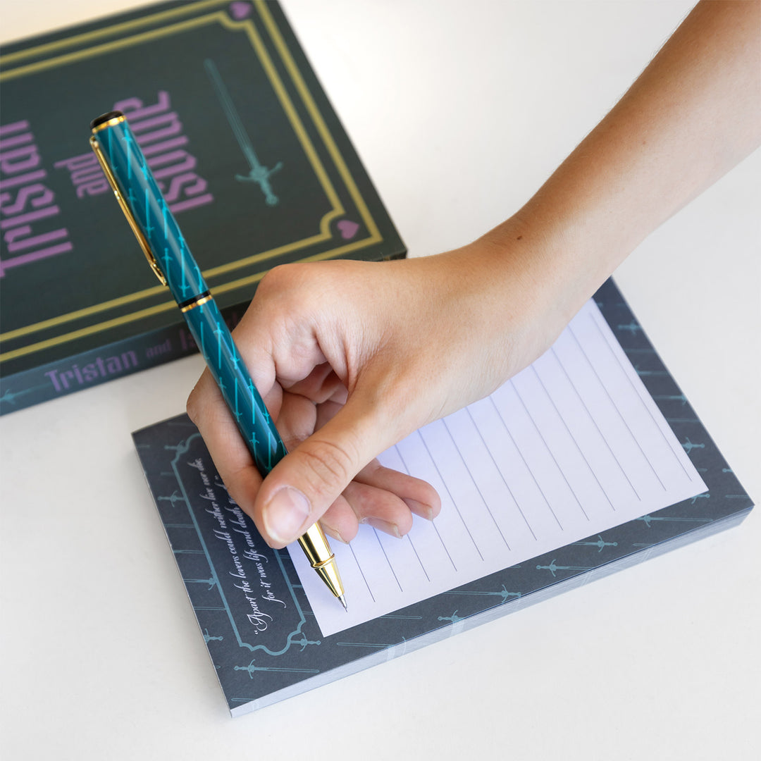 A hand holds a gold and blue pen with a light blue sword pattern on it over a lined notepad with matching background. Next to the notepad is a box labeled Tristan and Isolde.