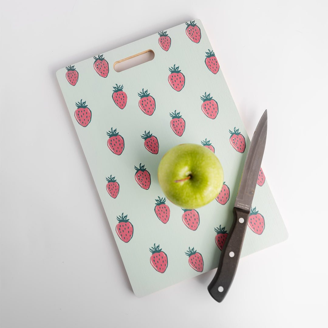 A strawberry-patterned cutting board with a green apple and a knife laying on top of it.
