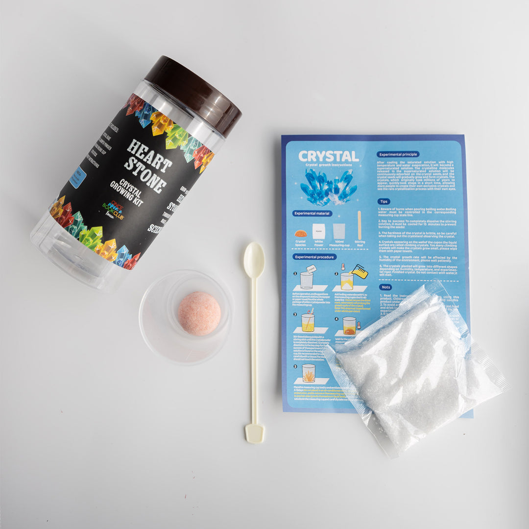 A Heart Stone Crystal Growing Kit jar next to the supplies included: instruction page, packet of crystals, spoon, and cup.