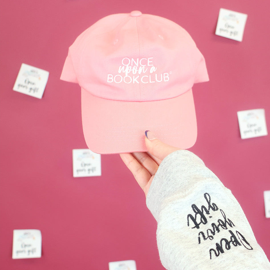 A pink baseball cap with 'Once Upon a book Club' embroidered in white on the front, held by a hand wearing a light gray sweatshirt with black sleeve embroidery that reads 'Open Your Gift' against a pink background with 'Open Your Gift' sticky notes.